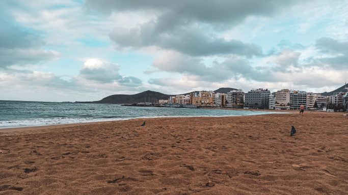 Strand mit feinem Sand, ruhigem Wasser und Küstenstadt im Hintergrund unter bewölktem Himmel.