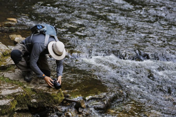 Person kniet am Ufer eines Flusses und fotografiert, während das Wasser fließt.